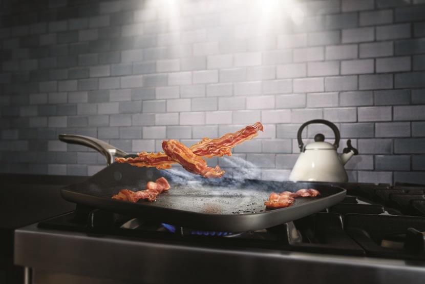 A stove top in kitchen with flying bacon over top a frying pan.